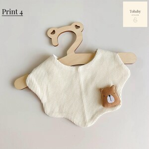 May include: A cream-colored waffle-textured bib with a scalloped edge, featuring a small brown bear applique on the lower right side. The bib is displayed on a light-colored wooden hanger. The text Print 4 and Tobaby Atelier are visible.