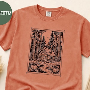 May include: A terracotta-colored short-sleeve t-shirt featuring a black ink-style graphic print of a small cottage nestled in a forest with pumpkins in the foreground. The shirt has a crew neckline and a Comfort Colors brand label at the inner neck.