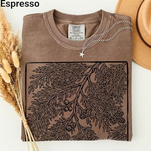 May include: Espresso-colored t-shirt with a black botanical print. The shirt features a crew neck and a Comfort Colors tag. A silver necklace with a star pendant is draped on the shirt. A tan hat is visible in the background.