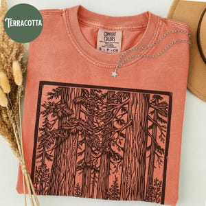 Może przedstawiać: T-shirt z kr&oacute;tkim rękawem w kolorze terakoty z czarnym nadrukiem graficznym przedstawiającym gęsty las z wysokimi drzewami i gałęziami. Koszulka ma okrągły dekolt i metkę Comfort Colors wewnątrz kołnierza. Na tkaninie spoczywa srebrny naszyjnik z gwiazdką.