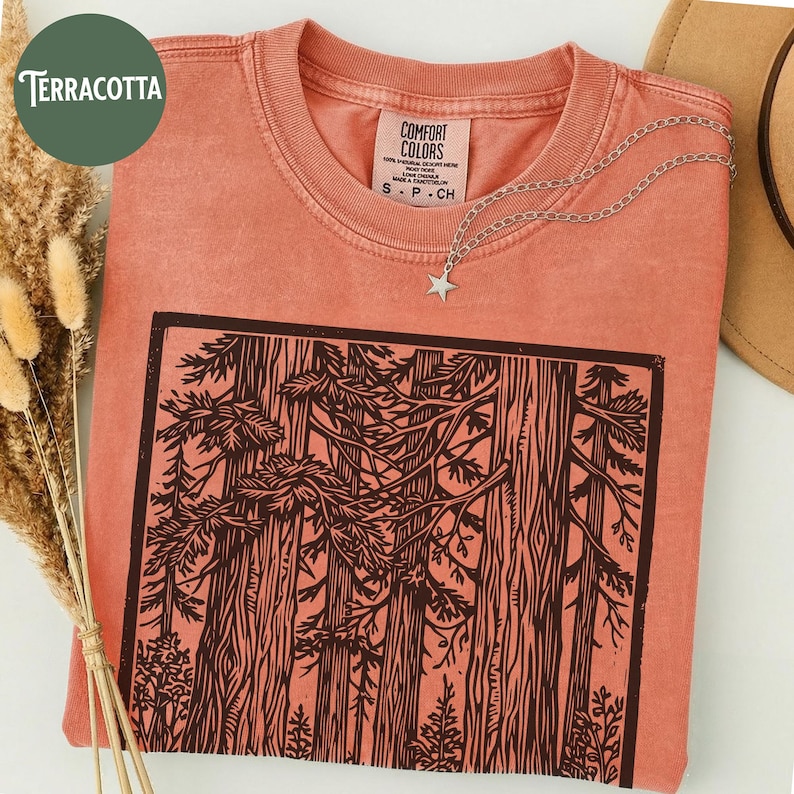 Może przedstawiać: T-shirt z kr&oacute;tkim rękawem w kolorze terakoty z czarnym nadrukiem graficznym przedstawiającym gęsty las z wysokimi drzewami i gałęziami. Koszulka ma okrągły dekolt i metkę Comfort Colors wewnątrz kołnierza. Na tkaninie spoczywa srebrny naszyjnik z gwiazdką.