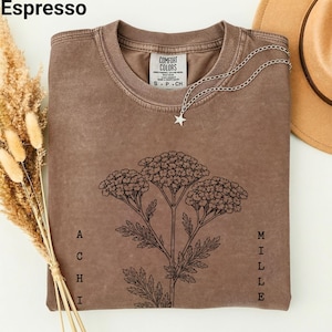 May include: Espresso-colored t-shirt with a floral graphic and the text "ACHILLE" and "MILLE". The shirt features a crew neck and a tag that reads "COMFORT COLORS". A silver necklace with a star pendant and a brown hat are also visible.