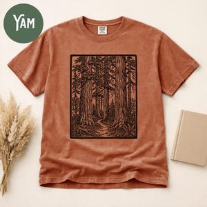 Może przedstawiać: T-shirt z kr&oacute;tkim rękawem w kolorze terakoty z czarnym nadrukiem graficznym w stylu tuszu, przedstawiającym gęsty las z wysokimi drzewami i krętą ścieżką. Koszulka ma okrągły dekolt i widoczną metkę marki wewnątrz obszaru kołnierza.