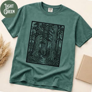 Może przedstawiać: Szałwiowy t-shirt z kr&oacute;tkim rękawem z czarnym, prostokątnym nadrukiem graficznym przedstawiającym gęsty las z wysokimi drzewami i krętą ścieżką. Koszulka ma okrągły dekolt i metkę marki Comfort Colors widoczną po wewnętrznej stronie kołnierza.
