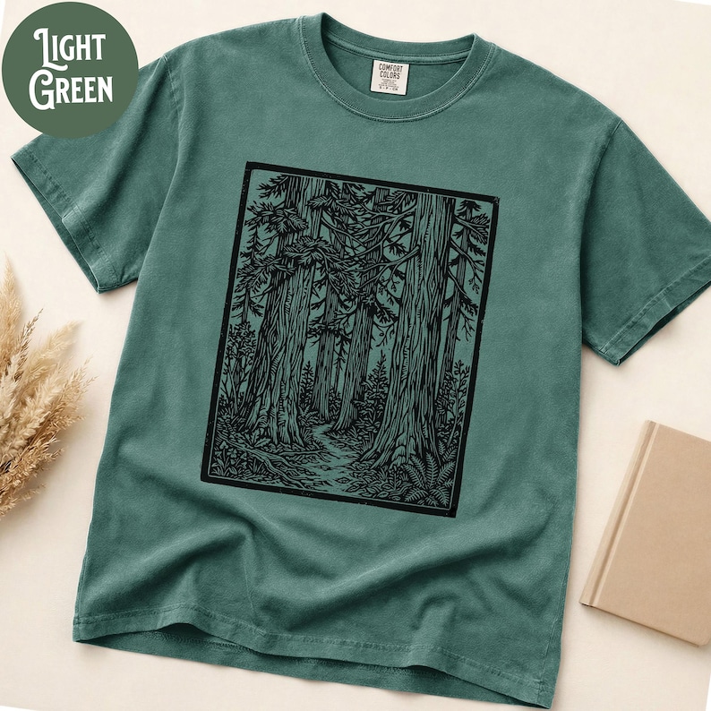 Może przedstawiać: Szałwiowy t-shirt z kr&oacute;tkim rękawem z czarnym, prostokątnym nadrukiem graficznym przedstawiającym gęsty las z wysokimi drzewami i krętą ścieżką. Koszulka ma okrągły dekolt i metkę marki Comfort Colors widoczną po wewnętrznej stronie kołnierza.
