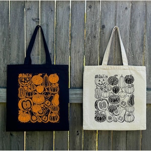May include: A black tote bag with an orange pumpkin print and a natural canvas tote bag with a black pumpkin print. Both bags feature two shoulder straps and a rectangular graphic design composed of various pumpkin and gourd illustrations.