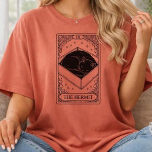 May include: A terracotta-colored t-shirt featuring a tarot card design. The card depicts a black cat curled up inside a rectangular frame, with the words "THE HERMIT" at the bottom. The card is framed with decorative elements.