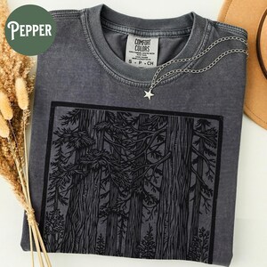 Może przedstawiać: Ciemnoszary t-shirt z okrągłym dekoltem i czarnym nadrukiem graficznym przedstawiającym gęsty las z wysokimi drzewami. Koszulka posiada metkę Comfort Colors przy kołnierzu i jest zestawiona z naszyjnikiem z srebrną zawieszką w kształcie gwiazdy.