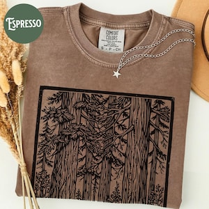 Może przedstawiać: Brązowy t-shirt z okrągłym dekoltem i czarnym nadrukiem graficznym przedstawiającym gęsty las z wysokimi drzewami w prostokątnej ramce. Koszulka posiada metkę Comfort Colors przy wewnętrznym dekolcie oraz srebrny naszyjnik z zawieszką w kształcie gwiazdy.