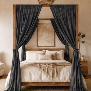 May include: A wooden four-poster bed frame featuring long, dark charcoal grey fabric curtains draped from the top rails and tied at the sides. The bed is dressed with white pillows, a beige lumbar pillow, and a light tan textured throw blanket.
