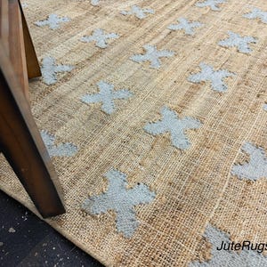 Può includere: Un tappeto in juta color marrone chiaro intrecciato con un motivo ripetitivo di croci azzurre. Il tappeto presenta una struttura in fibra naturale testurizzata con cuciture visibili. Il testo JuteRugsAura è stampato nell'angolo in basso a destra.