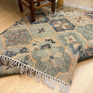 May include: A rectangular woven area rug featuring a repeating geometric pattern in shades of blue, sage green, and tan. The rug has a textured surface with a visible jute-like weave and white fringe along the edges. The text JuteRugsAura is visible.