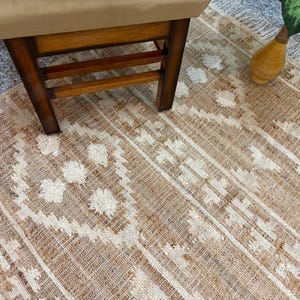 Peut inclure: Un tapis tissé présentant une palette de couleurs beige et crème avec un motif géométrique. Le tapis a une surface texturée avec des détails touffetés en relief de couleur crème, disposés en motifs de losanges et linéaires sur la base beige.