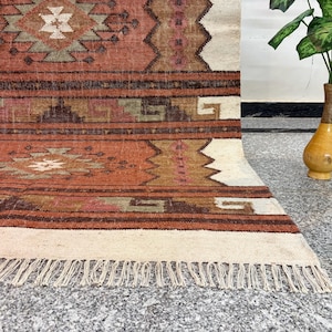 May include: A woven area rug featuring a geometric pattern in shades of terracotta, brown, cream, and muted green. The rug displays horizontal bands with intricate diamond and stepped motifs, finished with a cream-colored fringe along the bottom edge.