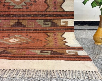 May include: A woven area rug featuring a geometric pattern in shades of terracotta, brown, cream, and muted green. The rug displays horizontal bands with intricate diamond and stepped motifs, finished with a cream-colored fringe along the bottom edge.