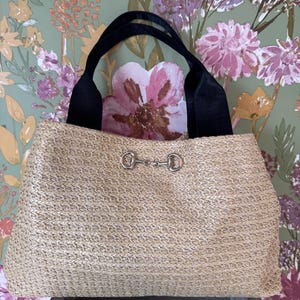 May include: A woven straw tote bag with black handles and silver-coloured metal hardware. The bag has a textured, natural-toned body and a rectangular shape. The handles are made of a smooth, black material.