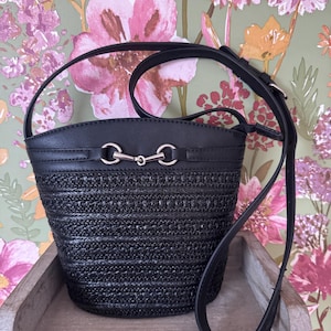 May include: Black woven bucket bag with a leather top and strap. The bag features silver-tone hardware and a long adjustable strap for shoulder or crossbody wear. The bag is sitting on a wooden tray with a floral wallpaper background.
