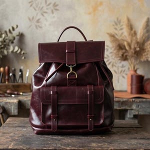 May include: A deep burgundy leather backpack featuring a fold-over top flap with a brass-toned metal hook closure, a top handle, and a large front pocket secured by two vertical leather straps with metal buckles and studs.