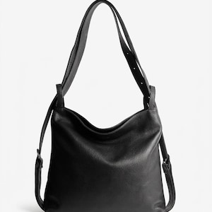 May include: A black leather hobo bag with a pebbled texture, featuring a slouchy silhouette, a single wide shoulder strap, and thin side straps with adjustable buckles. The bag has a curved top opening and a structured base for stability.