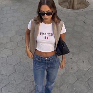 以下が含まれることがあります： 「FRANCE」とプリントされた白いクロップトップ、青いジーンズ、ベージュのベストを着た女性。黒いサングラスと黒いハンドバッグを身に着けています。カジュアルでスタイリッシュな服装です。