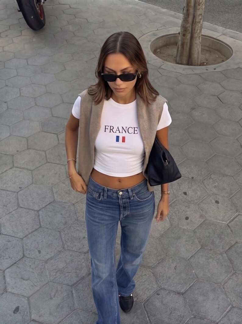 以下が含まれることがあります： 「FRANCE」とプリントされた白いクロップトップ、青いジーンズ、ベージュのベストを着た女性。黒いサングラスと黒いハンドバッグを身に着けています。カジュアルでスタイリッシュな服装です。