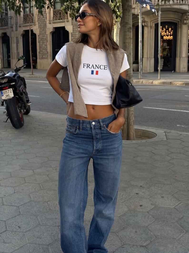 以下が含まれることがあります： 「FRANCE」の文字とフランス国旗がプリントされた白いクロップド丈トップスに、青いジーンズを合わせた女性。ベージュのセーターが肩にかけられています。サングラスと黒いバッグがアクセント。背景にはオートバイがあります。