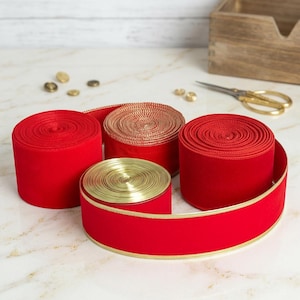 Op de afbeelding: Verschillende rollen rood fluwelen lint met gouden rand en een rol goudkleurig lint. Gouden schaar en kleine gouden versieringen zijn ook aanwezig. De linten zijn gerangschikt op een marmeren oppervlak, wat wijst op een knutsel- of cadeauverpakkingsthema.