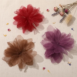 May include: Three layered fabric flowers in shades of red, brown, and purple. The flowers are arranged on a light-colored surface with dried flowers and petals scattered around them. The flowers have a delicate, airy appearance.