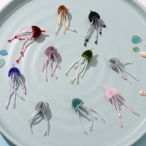 Puede incluir: Una colección de adornos de medusas con cuentas en varios colores, incluyendo rojo, marrón, negro, morado, verde, gris, azul y rosa. Cada medusa tiene una tapa texturizada y delicados tentáculos de cuentas. Los adornos están dispuestos sobre una superficie azul claro.