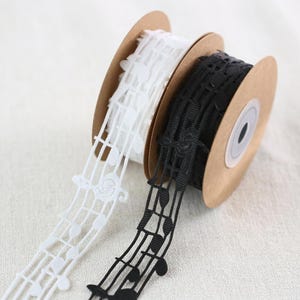 Puede incluir: Dos carretes de cinta decorativa, uno blanco y otro negro. Ambos presentan un diseño de notas musicales. Las cintas están en carretes de cartón marrón, ideales para manualidades, costura o envolver regalos.