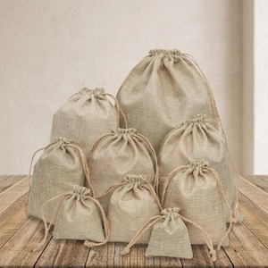 May include: A collection of eight beige drawstring pouches made of woven fabric, arranged in a tiered formation from smallest to largest. Each pouch features a simple gathered top secured with matching thin cord ties, showcasing a natural texture.