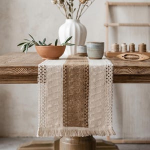 Può includere: Una tovaglia tessuta con una striscia centrale marrone e lati color crema. La tovaglia ha un bordo con frange ed è posizionata su un tavolo di legno. Articoli decorativi come un vaso, ciotole e tazze.