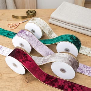 Könnte beinhalten: Rollen aus Samtband in verschiedenen Farben, darunter Burgunderrot, Lavendel und Smaragdgrün, mit goldenem Rand. Ein Maßband und eine Schere sind ebenfalls zu sehen, was auf Basteln hindeutet.