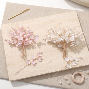 以下が含まれることがあります： ピンクと白の2つのフローラルヘアアクセサリー。繊細なビーズの花とワイヤーの茎が付いています。ベージュの表面に、金の針と糸巻きと一緒に配置されています。