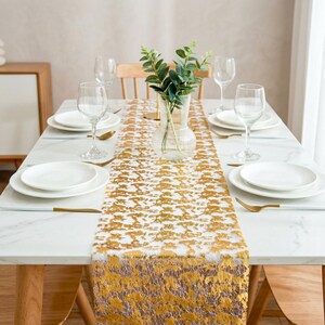 May include: A white marble dining table set for four with a gold metallic patterned table runner. The table features four white ceramic plates, four clear glass wine glasses, four gold forks, and a clear glass vase containing green eucalyptus.