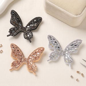 Pode incluir: Três enfeites em forma de borboleta com detalhes brilhantes semelhantes a cristais. Um é preto, outro ouro rosa e o outro prateado. As asas são intrincadamente detalhadas e as borboletas estão dispostas sobre uma superfície clara.