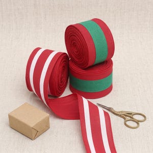 Può includere: Tre rotoli di nastro in tessuto. Un rotolo presenta uno sfondo rosso con due strisce bianche. Due rotoli presentano uno sfondo rosso con un'ampia striscia centrale verde. Una piccola scatola rettangolare marrone e forbici in metallo dorato.