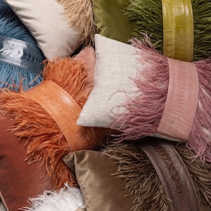 Pode incluir: Uma coleção de almofadas decorativas em várias cores e texturas. As almofadas apresentam veludo, linho e acabamentos em penas de avestruz sintéticas. Algumas almofadas têm faixas de couro ou couro sintético. As cores incluem creme, rosa, laranja, castanho, verde e azul.