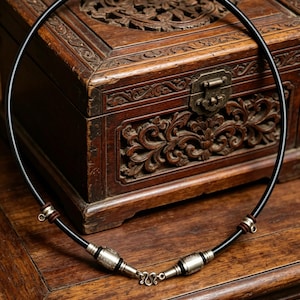 Puede incluir: Un collar negro con detalles metálicos plateados y un cierre. El collar se exhibe sobre una superficie de madera con intrincadas tallas, mostrando una estética vintage. El collar tiene forma circular.