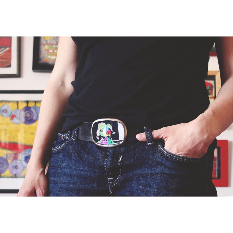 May include: A silver belt buckle with a colorful abstract design of a person's face. The buckle is attached to a black leather belt and worn over blue jeans.