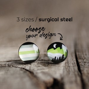 May include: Two round stud earrings with black rims. One features a striped pattern in black, white, and green. The other depicts a mountain scene with a crescent moon, green hills, and silhouetted trees. Text reads "3 sizes / surgical steel" and "choose your design".