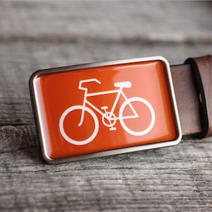 Peut inclure: Une ceinture en cuir marron avec une boucle argent&eacute;e repr&eacute;sentant une silhouette de v&eacute;lo blanche sur fond orange.