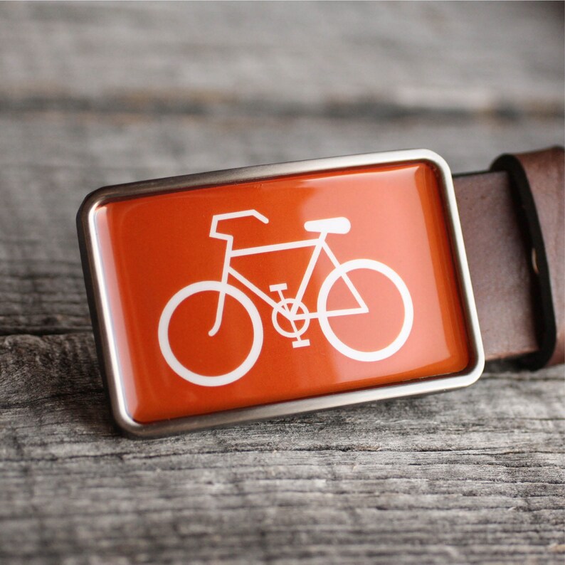 Peut inclure: Une ceinture en cuir marron avec une boucle argent&eacute;e repr&eacute;sentant une silhouette de v&eacute;lo blanche sur fond orange.