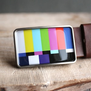May include: A silver belt buckle featuring a colourful TV test pattern design. The pattern includes red, orange, yellow, green, blue, purple and black.