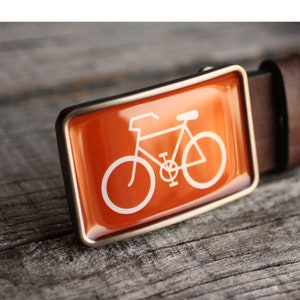 Peut inclure: Une boucle de ceinture argent&eacute;e avec un fond orange et une silhouette blanche d'un v&eacute;lo.