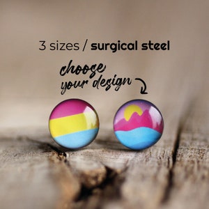 Puede incluir: Dos pendientes redondos de cristal con un respaldo plateado. El pendiente izquierdo presenta un diseño rosa, amarillo y azul. El pendiente derecho presenta un diseño de puesta de sol rosa, amarillo y azul. El texto "choose your design" está escrito encima de los pendientes.