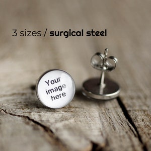Könnte beinhalten: Nahaufnahme von zwei runden Ohrsteckern mit silbernem Stecker und Verschluss. Die Ohrringoberfläche ist weiß mit dem Text "Your image here". Der Text "3 sizes / surgical steel" befindet sich über den Ohrringen.
