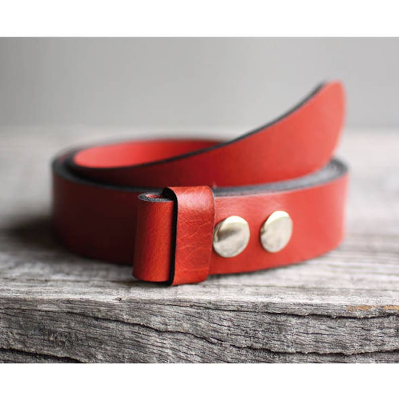 Op de afbeelding: Een opgerolde rode leren riem met zilverkleurige klinknagels. De riem heeft een levendige rode kleur en lijkt gemaakt van hoogwaardig leer. De riem ligt op een verweerd houten oppervlak, wat de textuur en het ontwerp laat zien.