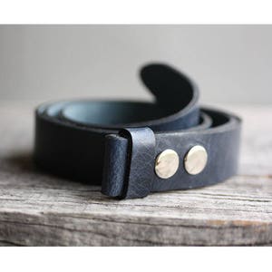 Op de afbeelding: Een donkerblauwe leren riem met een zilveren metalen gesp en twee zilveren metalen klinknagels. De riem is opgerold en de gesp is gericht op de camera.