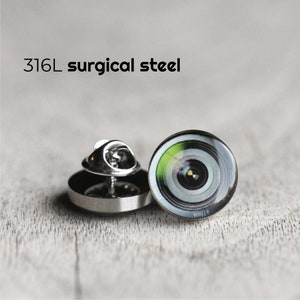 Könnte beinhalten: Eine silberne Anstecknadel mit einem Schwarzweißbild eines Kameraobjektivs. Die Nadel ist aus 316L-Chirurgenstahl.